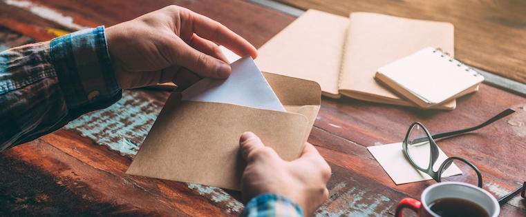 Close-up of hands opening a kraft paper envelope at a wooden table with notebooks, glasses, and a coffee cup; warm rust and beige mood